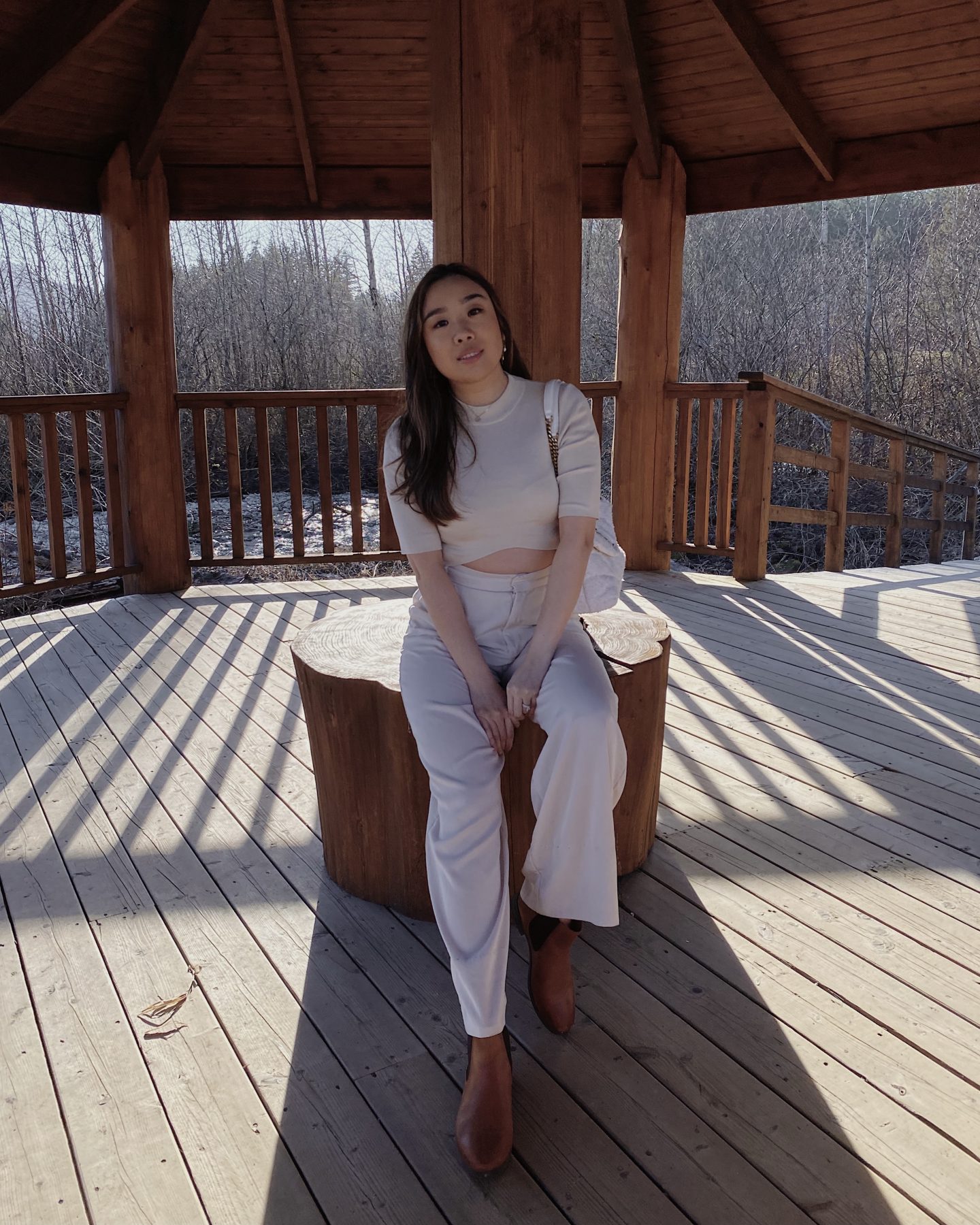 outdoor background on wooden patio. Woman sitting on wooden stump with white top and white pants