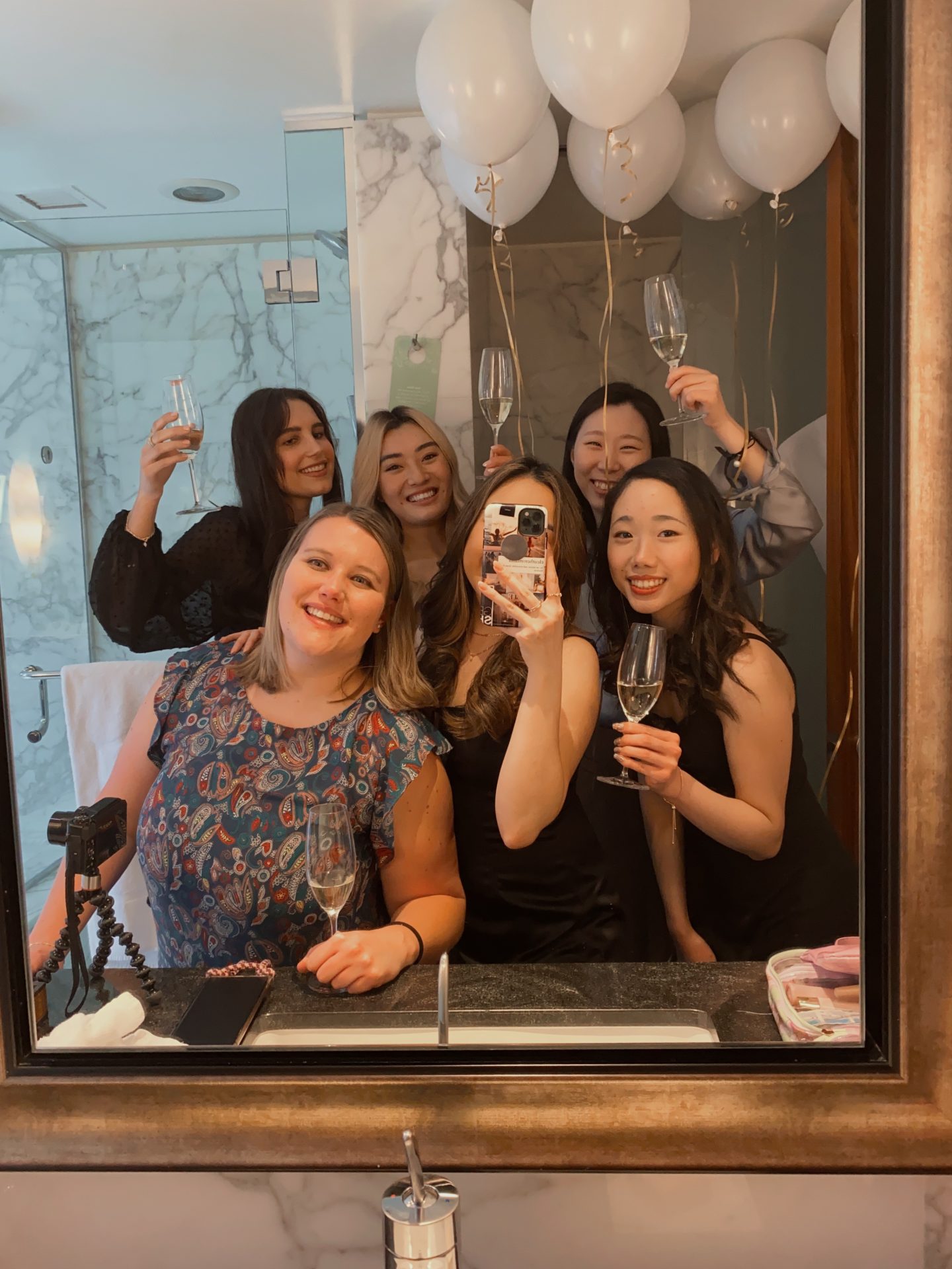 6 ladies posing in a mirror holding glasses with balloons in the background